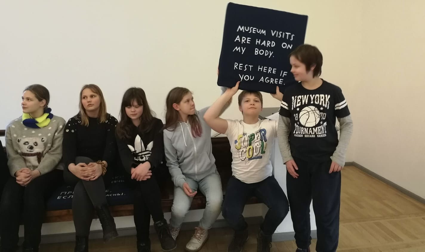 Kuus õpilast istumas-seismas Tallinna Kunstihoone näitusel, ühel poisil on käes must padi valge tekstiga "Museum visits are hard on my body. Rest here if you agree."
