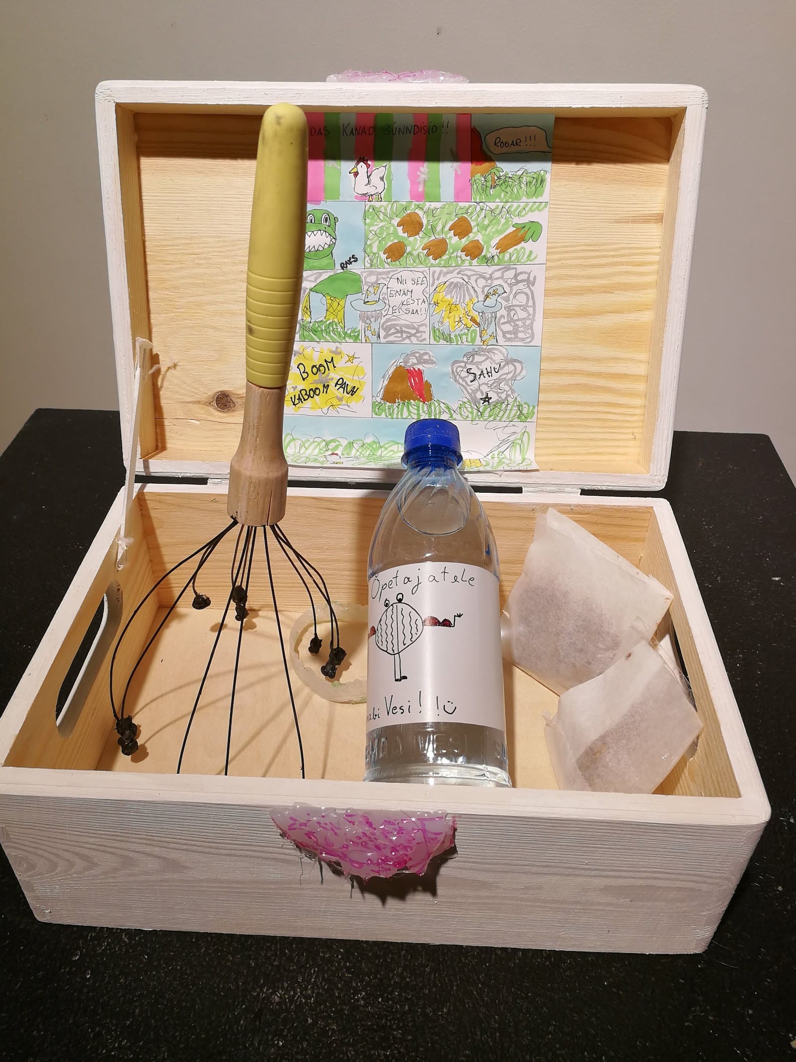 Valgeks peitsitud puidust karp, mille sisekaanel on tuju tõstev koomiks ning sees esmaabi vahendid õpetajatele - pudel vett, küpised ja peamassaaži seadeldis
