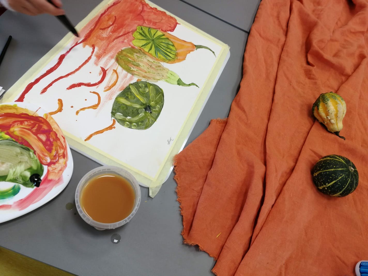 Fotol on kujutatud oranži draperii ja minikõrvitsatega seadeldis ja õpilase käsi, kes seda parasjagu maalib koos pooliku maaliga